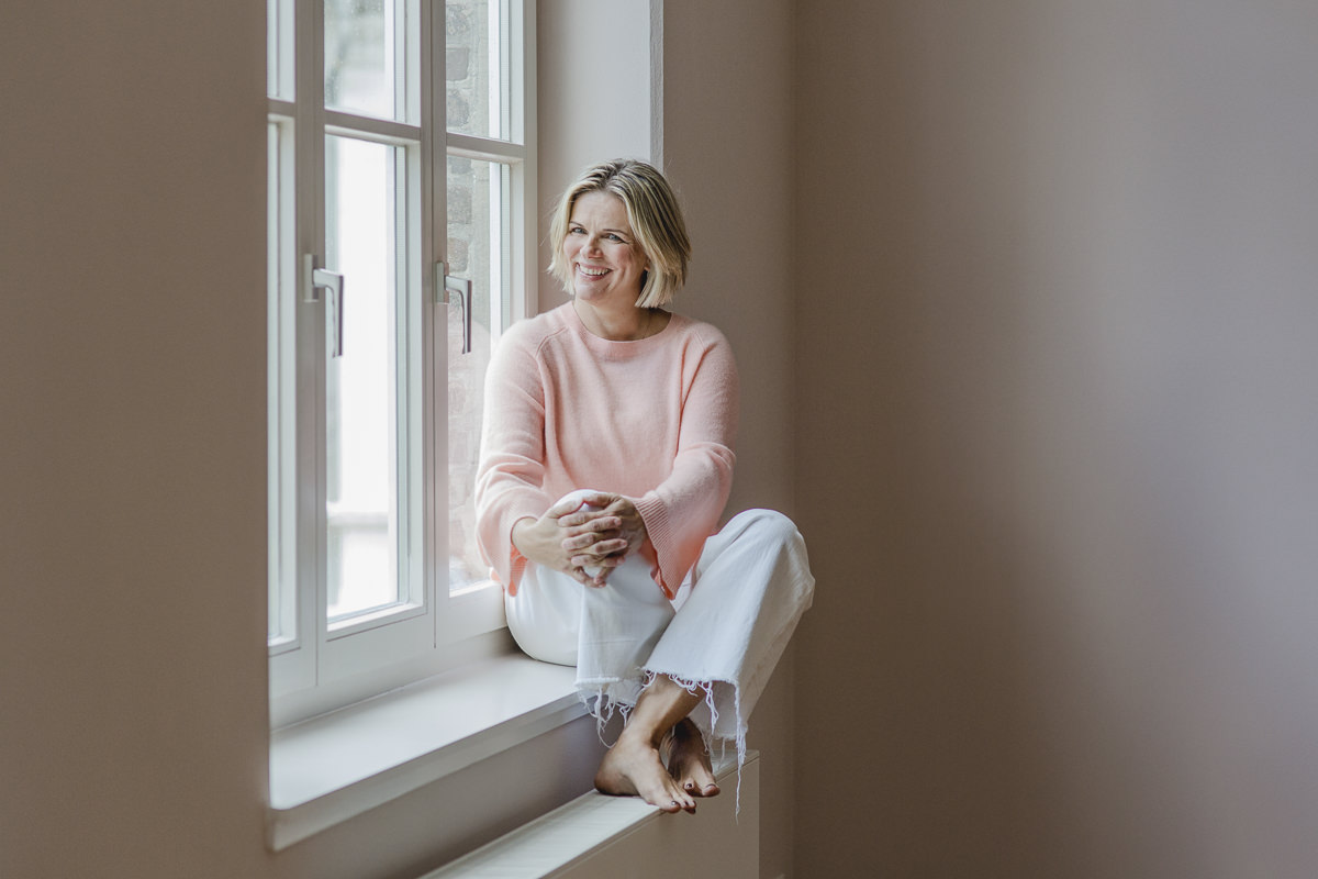 Portraitfoto von Yogatherapeutin Daniela Stilke, die lächelnd an einem Fenster sitzt