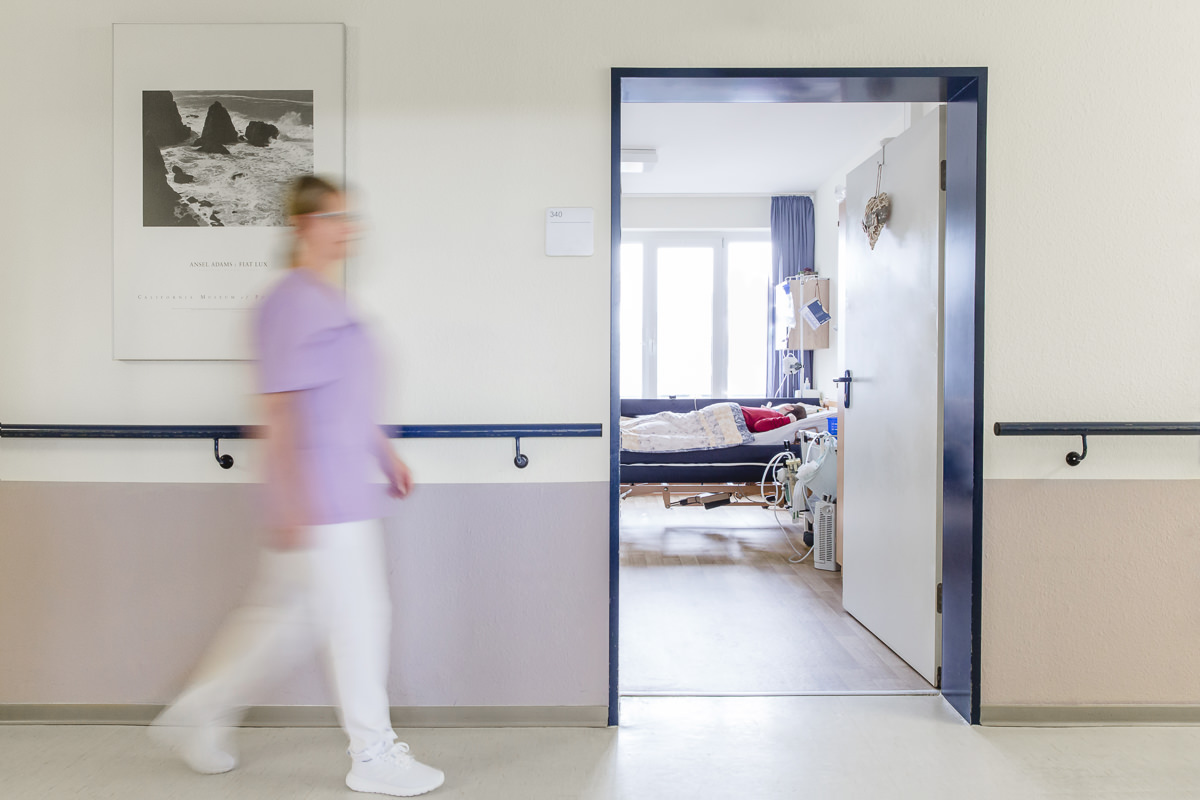 Blick in das Zimmer eines Langzeitpflege Patienten, während auf dem Flur eine Pflegerin vorbeigeht | Foto: Hanna Witte