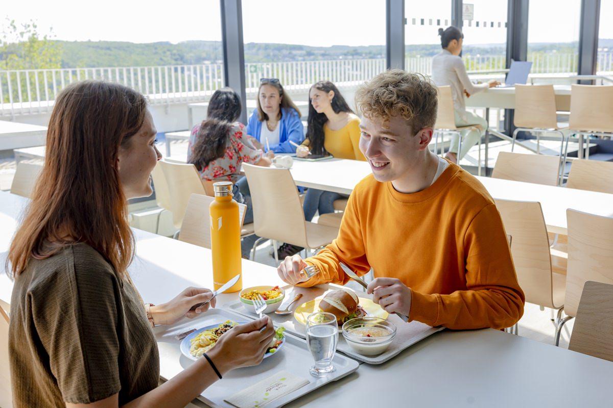 Studenten der Uni Siegen sitzen an Tischen der Cafeteria, essen und unterhalten sich | Foto: Hanna Witte