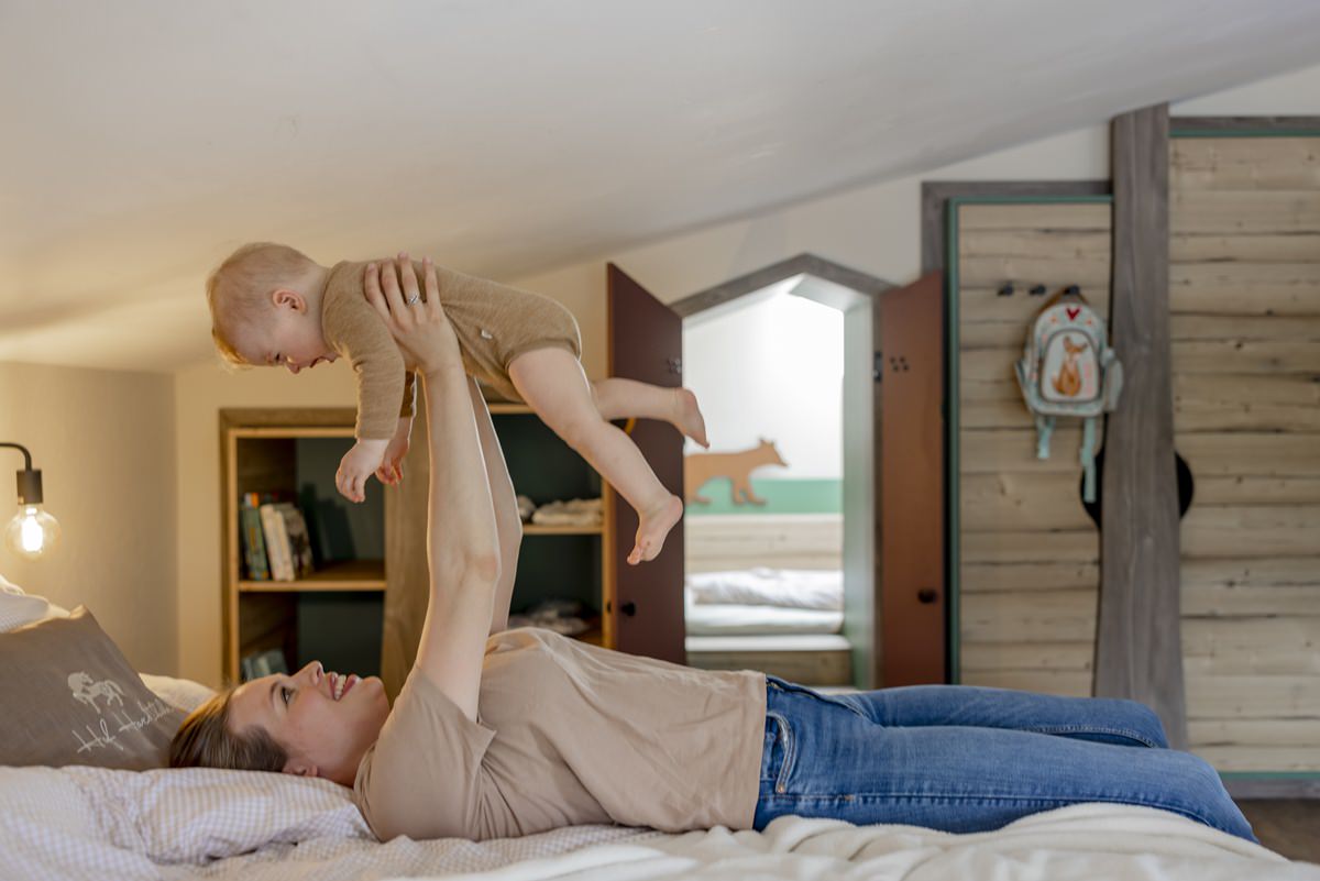eine Mutter liegt auf dem Bett ihres Ferienhauses und hebt glücklich ihr Baby in die Luft | Foto: Hanna Witte
