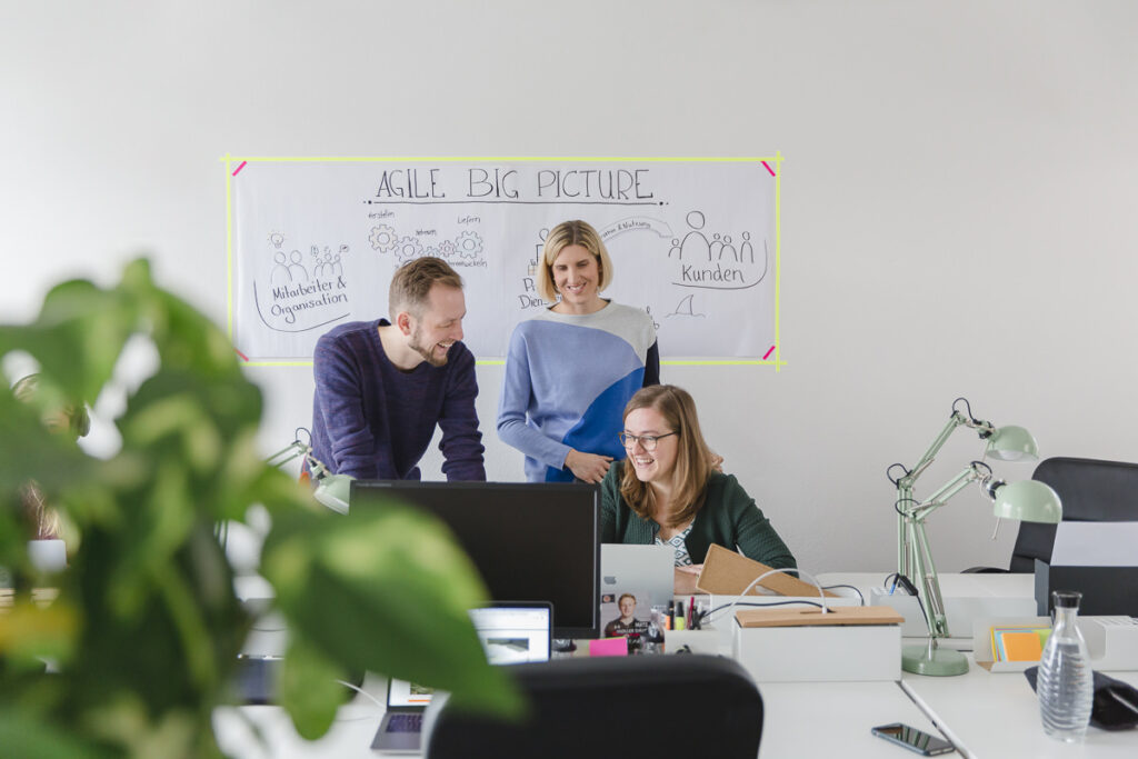 drei MAERA Mitarbeiter schauen auf einen Computer, dahinter hängt als Hintergrund ein Poster mit Arbeitsschritten | Foto: Hanna Witte